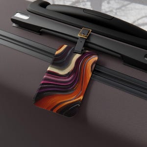May include: Luggage tag with a colorful abstract design. The tag features a black strap and buckle, and the design includes swirls of orange, purple, white, and black. The tag is attached to a dark gray suitcase.