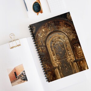 May include: Spiral-bound notebook with a detailed, golden steampunk design. The cover depicts an arched doorway with intricate clockwork gears. The notebook is open on a white surface, with a gold paper clip and sunglasses nearby.