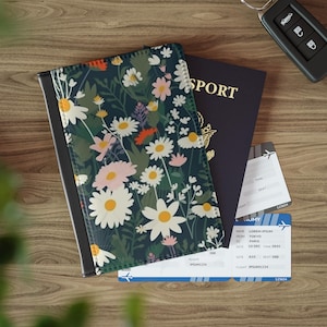 Könnte beinhalten: Eine Reisepasshülle mit dunkelblauem Hintergrund und einem wiederkehrenden Muster aus weißen Gänseblümchen, rosa Blumen und grünem Blattwerk. Die Hülle liegt auf einem dunkelblauen Reisepass und zwei weißen Bordkarten mit blauen Akzenten.