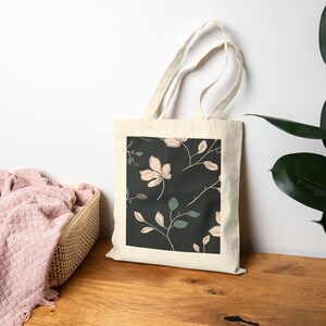 May include: A cream-colored tote bag with a floral pattern. The pattern features light pink flowers and green leaves on a dark background. The bag is sitting on a wooden surface.