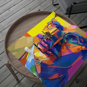 May include: A wrapped gift with a brown ribbon sits on colourful wrapping paper featuring a pop art portrait. A pair of scissors and a roll of tape are also on the paper. The scene is set on a wooden table.