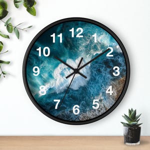 May include: A round wall clock with a black frame and a face featuring an aerial photograph of blue ocean waves crashing against a shoreline. The clock has white sans-serif numerals from 1 to 12 and black hour, minute, and second hands.