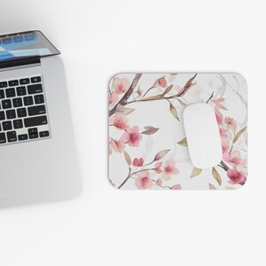 May include: A rectangular mousepad with a floral design featuring pink cherry blossoms and brown branches on a white background. A white computer mouse rests on the pad, next to a laptop.