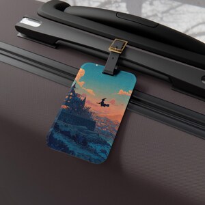 May include: A rectangular luggage tag with a vibrant illustration of a witch flying over a town at sunset. The tag has a black strap with a gold-coloured buckle, attached to a dark grey suitcase.