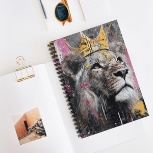 May include: Spiral-bound notebook featuring a regal lion wearing a gold crown. The lion's portrait is rendered in shades of gray, with pink and yellow paint strokes adding a pop of color. The notebook is open on a white surface.