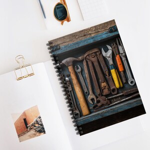 May include: Spiral-bound notebook featuring a detailed image of vintage tools arranged in a blue wooden toolbox. The tools include a hammer, wrenches, pliers, and other metal instruments, all with a rustic, aged appearance. The notebook is open on a white surface.