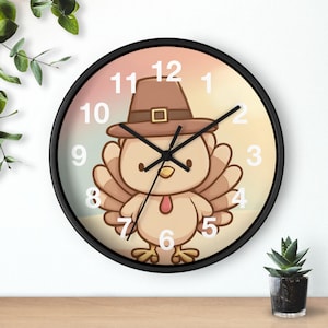 May include: A round wall clock with a black frame. The clock face features a cartoon turkey wearing a pilgrim hat. The numbers are white, and the hands are black.