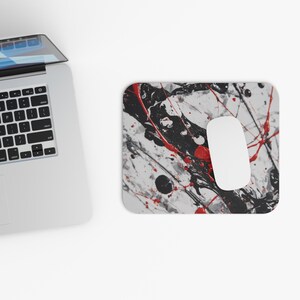Puede incluir: Un ratón de ordenador blanco descansa sobre una alfombrilla rectangular con un diseño abstracto de salpicaduras en negro, rojo y blanco. Un ordenador portátil plateado es parcialmente visible en la esquina superior izquierda.