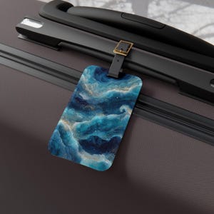 May include: A rectangular luggage tag with a blue and gold abstract design, attached to a black suitcase handle. The tag features a swirling pattern of blues and gold, resembling clouds or ocean waves. The tag is secured with a black strap and a gold-colored buckle.