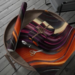 May include: A roll of wrapping paper and a gift box wrapped in the same paper, featuring a fluid, abstract pattern of swirling orange, deep purple, black, and cream-colored lines. A pair of silver scissors lies on the paper surface.