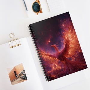 May include: Spiral-bound notebook featuring a vibrant illustration of a phoenix rising from flames. The artwork showcases a fiery color palette of red, orange, and purple hues, with the bird's wings spread wide against a dark background.