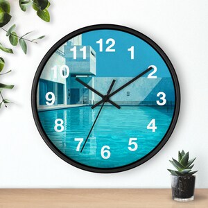 May include: A round wall clock with a black frame. The clock face features a blue pool scene with white architectural elements and black clock hands. White numbers mark the hours.