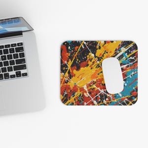 Puede incluir: Una alfombrilla de ratón rectangular con un ratón blanco y un diseño abstracto colorido. La alfombrilla presenta un fondo negro con salpicaduras de pintura amarilla, naranja, roja, blanca y azul. Un ordenador portátil es parcialmente visible en la esquina superior izquierda.