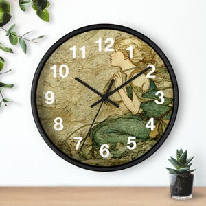 May include: A round wall clock with a black frame. The clock face features an illustration of a woman with long blonde hair in a flowing green dress. White numerals mark the hours. The clock hands are black.