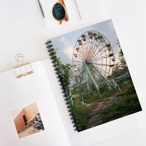 May include: Spiral-bound notebook featuring a photograph of a decaying Ferris wheel. The image shows a rusted, teal and red Ferris wheel surrounded by overgrown vegetation under a cloudy sky. The notebook is open on a white surface.