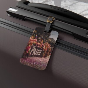 May include: A luggage tag with a black strap and buckle, displaying a blurred image of a path with the word "PRIDE" in white. The path is lined with pink and purple flowers. The tag is attached to a grey suitcase.