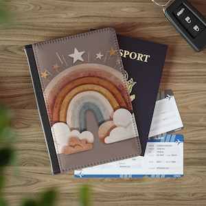 May include: A passport cover with a watercolor rainbow design, a passport, and two boarding passes are displayed on a wooden surface. The passport cover features a brown background with a rainbow, stars, and clouds. The passport is navy blue with gold lettering.