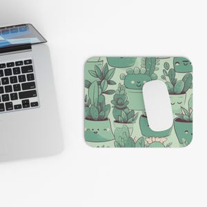 Op de afbeelding: Een rechthoekige muismat met een patroon van vetplanten in pot in groen- en wittinten. De muismat heeft een lichtgroene achtergrond en een witte computermuis ligt erop. Een laptop staat op de achtergrond.