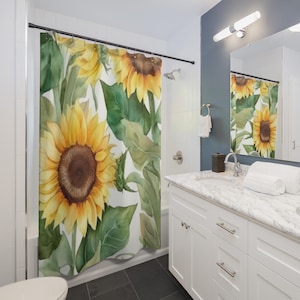 Pode incluir: Uma cortina de duche com um design em aquarela de grandes girassóis com pétalas amarelas e centros castanhos, rodeados por folhas verdes. A cortina está pendurada numa casa de banho com acessórios brancos e um chão escuro.