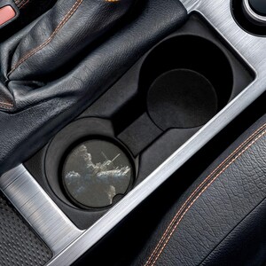 May include: A black car cup holder with a coaster featuring a cloudy sky design. The coaster is placed in the front cup holder. The interior of the car has black leather seats with orange stitching and silver trim.