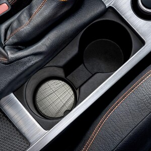 May include: A black car cup holder with a silver trim. The cup holder has two circular compartments and a textured coaster in the front. The surrounding interior features black leather and orange stitching.