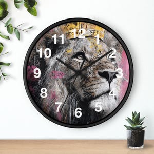 May include: Black-framed wall clock featuring a lion's face wearing a gold crown. The lion's portrait is in shades of gray, white, and brown, set against a pink background. White numerals mark the hours.