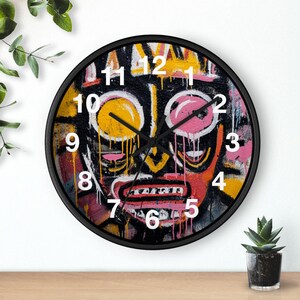 Puede incluir: Un reloj de pared con marco negro que presenta un colorido diseño de rostro abstracto. La cara tiene formas de ojos amarillos y rosas, una boca roja y números blancos. Las manecillas del reloj son negras.