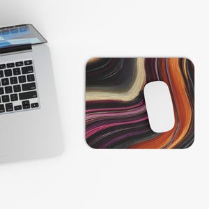 May include: A rectangular mouse pad featuring a swirling abstract pattern of black, orange, cream, and magenta lines. The mouse pad is positioned next to a silver laptop with a black keyboard and a white wireless computer mouse resting on top.