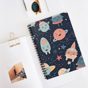 May include: Spiral-bound notebook with a dark blue cover featuring a whimsical space-themed pattern. The design includes smiling planets, stars, and other celestial bodies in shades of blue, pink, and yellow. The notebook is open, revealing blank pages.