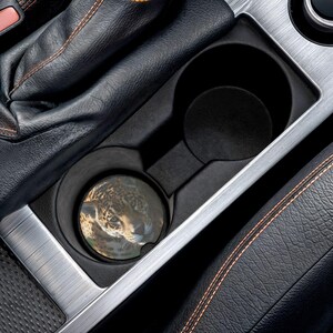 May include: A circular car cup holder coaster featuring a detailed photographic print of a jaguar face with characteristic black rosettes on a golden-brown coat. The coaster is positioned inside a black plastic vehicle center console cup holder.