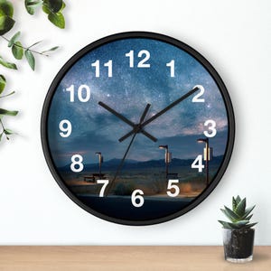 May include: A round wall clock with a black frame and a face featuring a night sky landscape with stars, mountains, and outdoor lighting. The clock has white Arabic numerals from 1 to 12 and black hour, minute, and second hands.