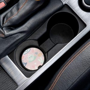 May include: A car cup holder with a pink coaster featuring a pattern of bunnies, teacups, and hearts. The coaster is round and fits snugly in the cup holder, which is black and part of a car's interior.