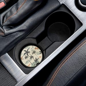 May include: A round car coaster with a tropical design featuring palm trees, airplanes, and other elements in shades of beige, green, and orange. The coaster is placed inside a car's cup holder.