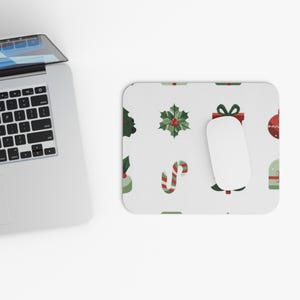 May include: A white rectangular mouse mat featuring a Christmas pattern with green holly, red baubles, gift boxes, and candy canes. A white computer mouse sits on the mat, next to a laptop.