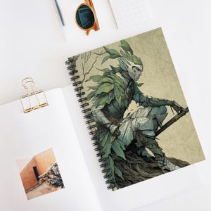 Puede incluir: Cuaderno de espiral con una ilustración de fantasía de una figura humanoide adornada con hojas verdes y armadura. La obra de arte utiliza verdes apagados, grises y beige. El cuaderno está abierto sobre una superficie blanca.