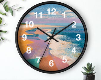 May include: A round wall clock with a black frame. The clock face features a colourful beach scene with shades of blue, orange, and purple. White numerals mark the hours, and black hands indicate the time. The clock is approximately 30 cm in diameter.