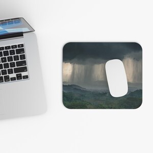 May include: A rectangular mousepad featuring a landscape scene of a rainforest under a dark, stormy sky with heavy rainfall. A white wireless mouse is positioned on the right side of the mousepad. A laptop is partially visible on the left.