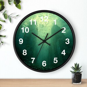 Puede incluir: Reloj de pared redondo con marco negro y escena submarina verde. La esfera del reloj tiene números blancos y manecillas negras. El fondo representa edificios sumergidos y peces.