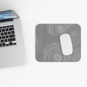 Op de afbeelding: Een grijze muismat met een wit wervelpatroon en een witte computermuis. De muismat is rechthoekig met afgeronde hoeken. Een zilveren laptop is ook zichtbaar.