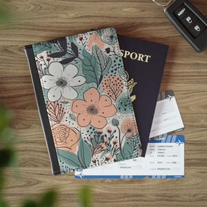 Op de afbeelding: Een paspoorthoes met een bloemenpatroon in perzik-, wit- en saliegroene tinten, die gedeeltelijk een donkerblauw paspoort met het woord PASSPORT in goud bedekt, samen met twee instapkaarten en een zwarte autosleutelhanger.