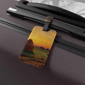 Può includere: Etichetta per bagagli con un dipinto di paesaggio di un campo con balle di fieno sotto un cielo al tramonto. L'etichetta è rettangolare con angoli arrotondati, attaccata a un cinturino nero con una fibbia color oro. L'etichetta è attaccata a una valigia grigio scuro.