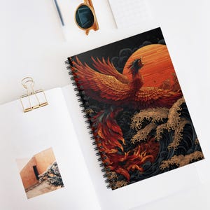 May include: Spiral-bound notebook featuring a vibrant illustration of a phoenix with outstretched wings against a backdrop of a setting sun and stylized waves. The color palette includes shades of red, orange, and black.