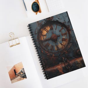 Pode incluir: Caderno espiralado com um design de mostrador de relógio desgastado. A capa apresenta um relógio enferrujado e envelhecido com algarismos romanos em tons de ouro e castanho sobre um fundo azul-acinzentado. O caderno está aberto, revelando páginas brancas.