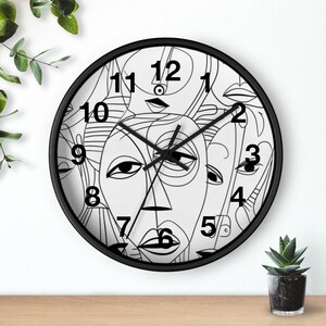 May include: A round wall clock with a black frame and a white face featuring a black line art pattern of multiple abstract faces. The clock has black hour, minute, and second hands, and large black numbers from 1 to 12 printed on the face.