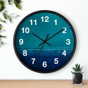 May include: A round wall clock with a black frame. The clock face features a gradient of teal and navy blue, with white numerals and black hands. The clock is displayed on a wooden surface with greenery in the background.