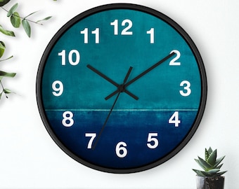May include: A round wall clock with a black frame. The clock face features a gradient of teal and navy blue, with white numerals and black hands. The clock is displayed on a wooden surface with greenery in the background.