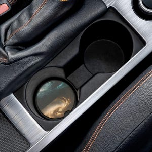 以下が含まれることがあります： シルバーのトリムが付いた黒い車のカップホルダー。渦巻く雲のデザインの丸いコースターがカップホルダーに入っています。車の内装は黒革とオレンジ色のステッチです。