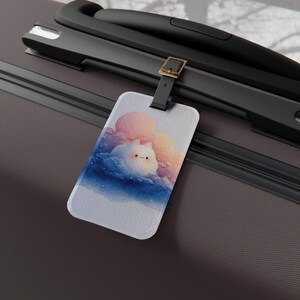 May include: A rectangular luggage tag with a black strap and gold-tone buckle attached to a dark gray suitcase. The tag features a printed illustration of a small white creature with a simple face resting among soft pink, blue, and purple clouds.