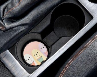 May include: A car coaster featuring a cartoon design of three Easter bunnies holding baskets. The coaster is round with a light pink background and is placed inside a car's cup holder. The car interior is black and silver.
