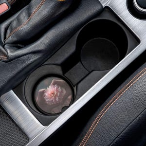 May include: A car cup holder with a floral car coaster. The coaster features a pink flower design with a blurred background. The cup holder is black and silver, and the car interior is black leather.